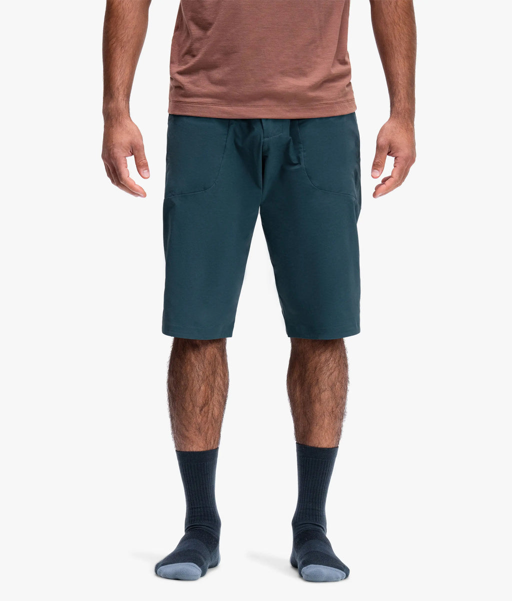A man wearing dark teal drawstring shorts, a dusty rose t-shirt, and dark socks, shown from the waist down against a light gray studio background.
