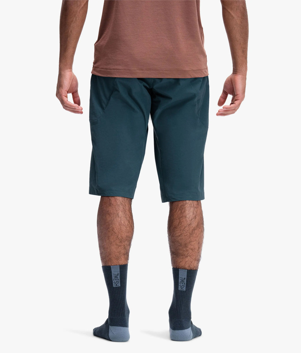 Rear view of a man wearing dark teal athletic shorts, a brown t-shirt, and dark gray socks against a light gray studio background.