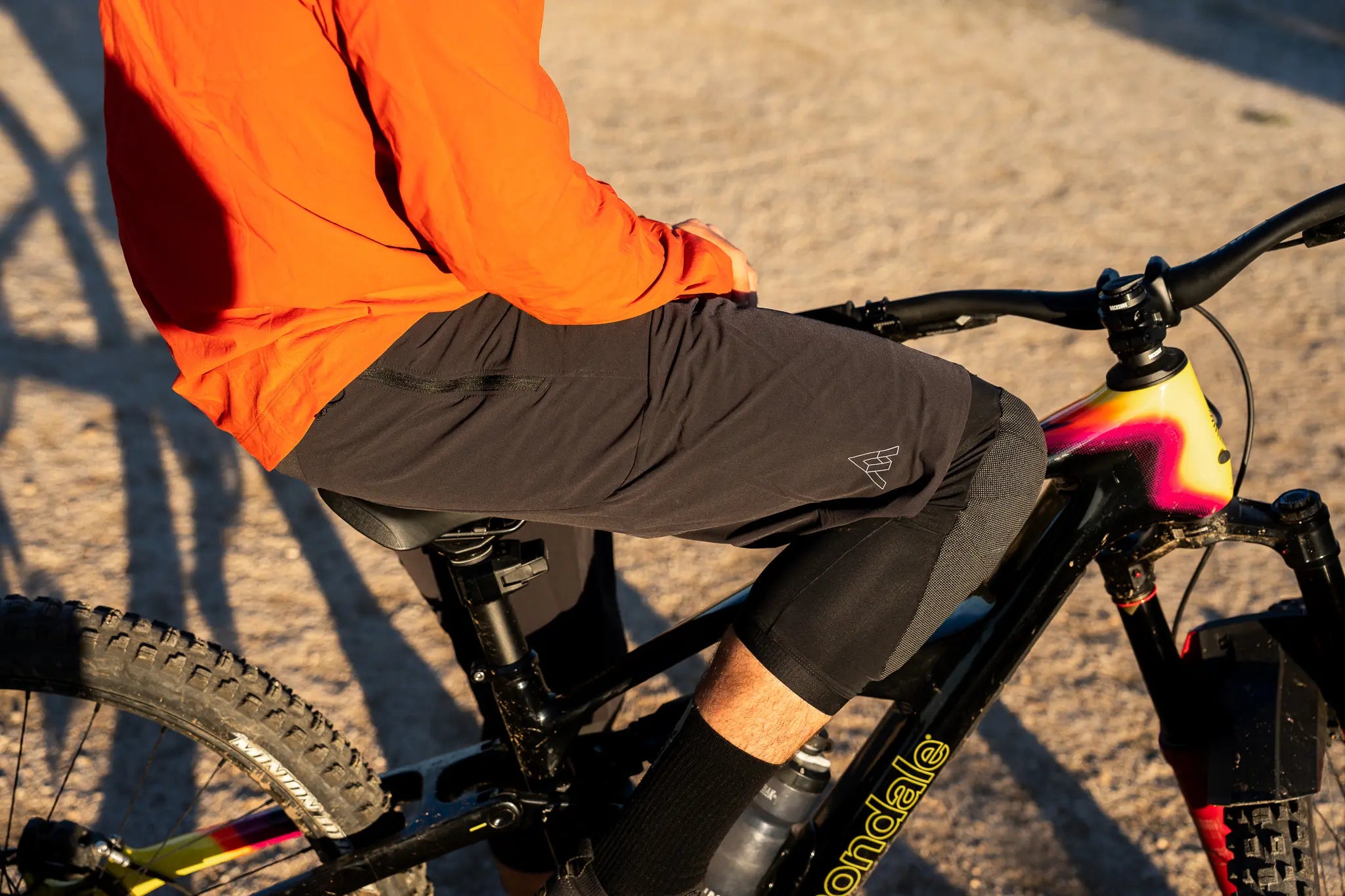 A close-up lifestyle shot of a person riding a mountain bike outdoors. The rider is wearing a bright orange long-sleeve top and dark gray athletic shorts, seated on a black mountain bike with a yellow and pink frame accent.