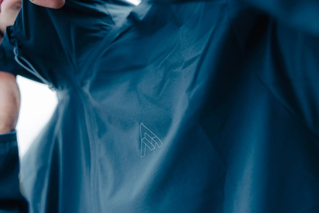 A close-up of a dark blue waterproof jacket being held open by a person, showing a subtle white outlined mountain logo on the chest.