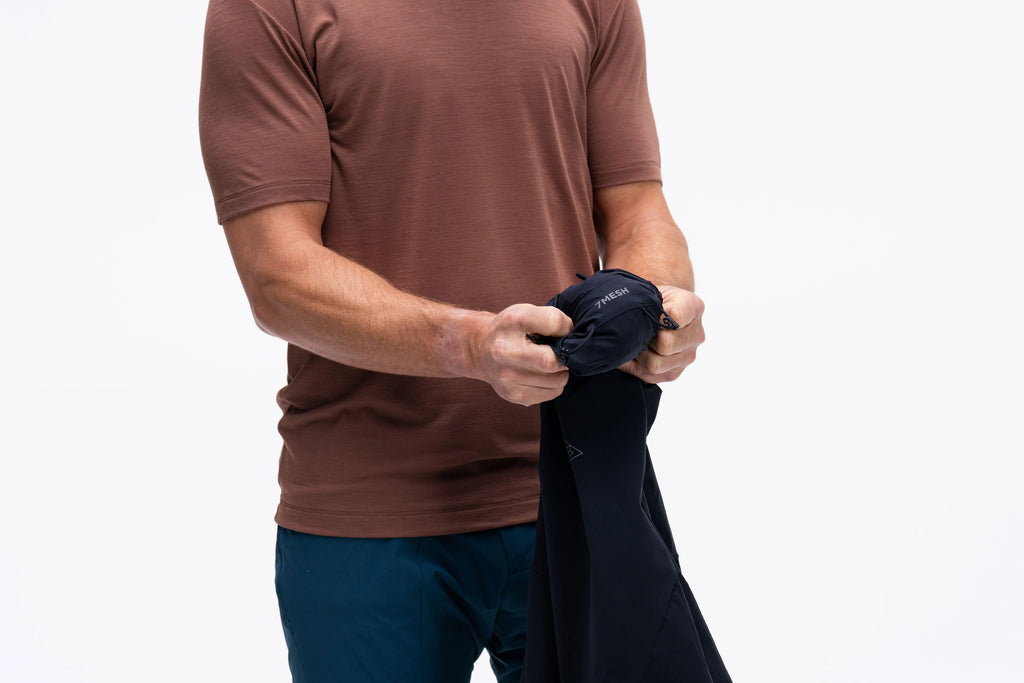 A man wearing a brown short-sleeve athletic shirt and dark blue pants is shown from the chest down, holding and stretching a black sleeveless undergarment or towel in his hands against a plain white background.