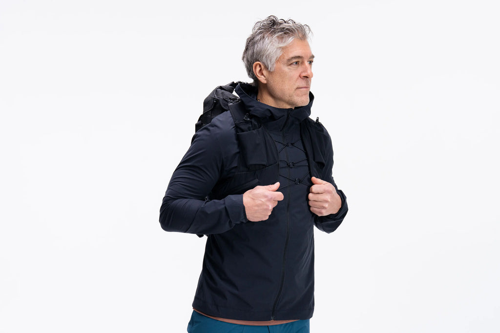 A man wearing a black hooded jacket, standing against a solid white background and looking to the side while holding the collar of the jacket.