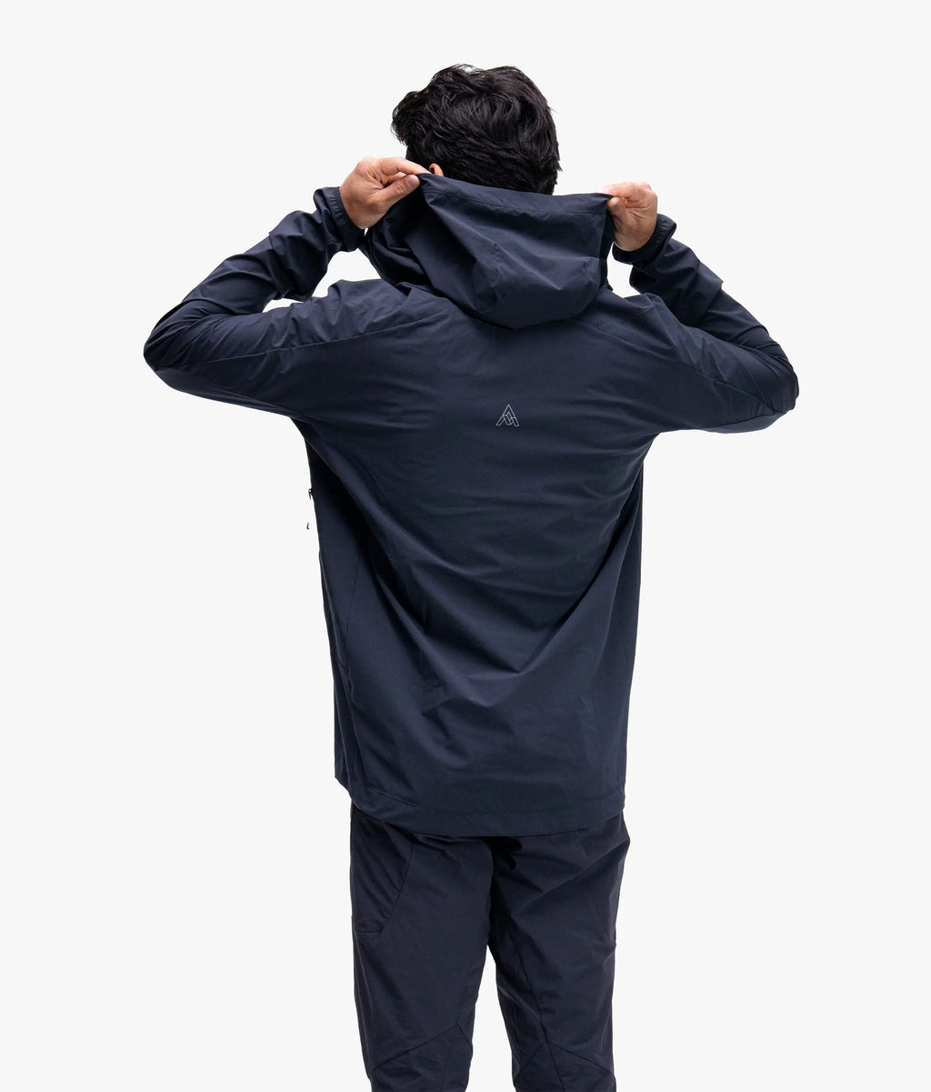 A man viewed from behind wearing a dark navy hooded jacket and matching pants, adjusting the hood against a plain light gray studio background.