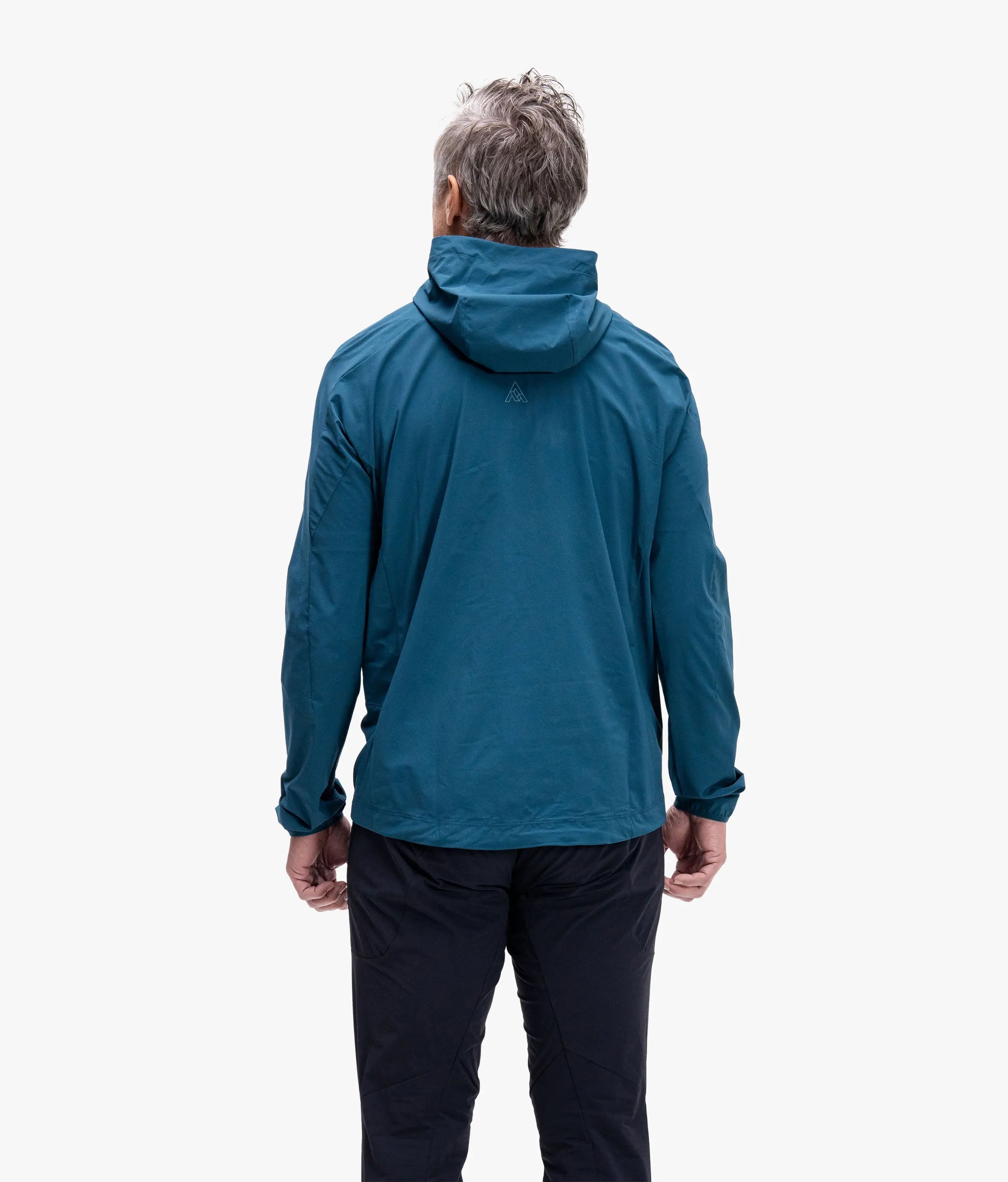 A man viewed from behind wearing a teal blue hooded jacket and dark pants against a plain white studio background.