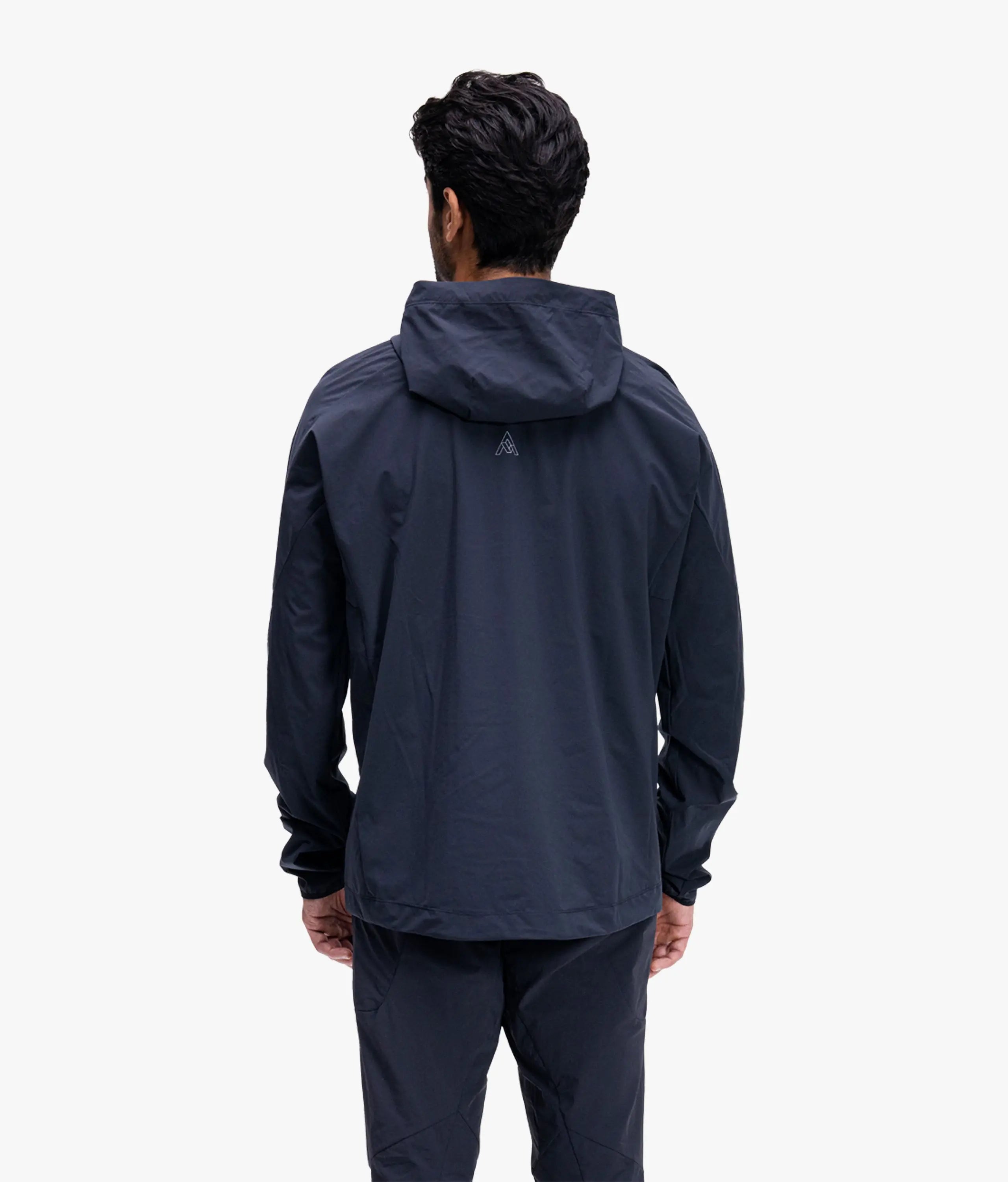 A man viewed from behind wearing a dark navy hooded jacket and matching dark pants against a clean white studio background.