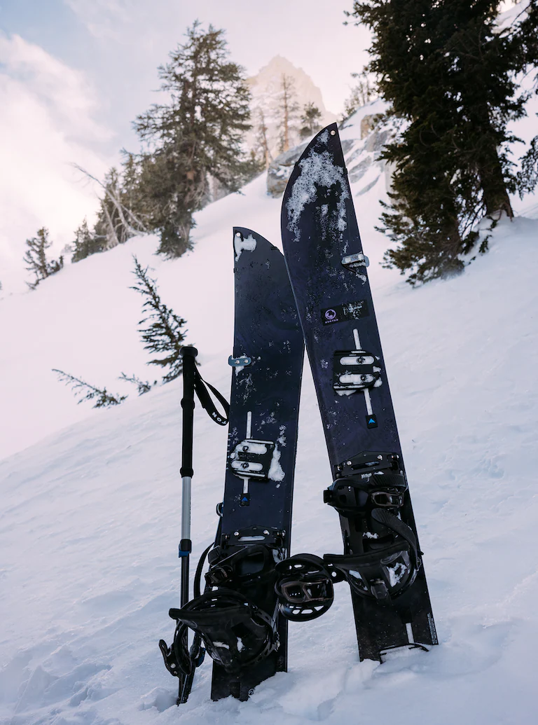 Two skis with bindings and a ski pole on a snowy slope with trees in the background.