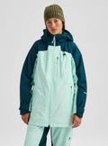 A model wearing a color-blocked snowboard jacket in mint green and dark teal, paired with matching pants, a striped beanie, and a face mask, against a light gray studio background.