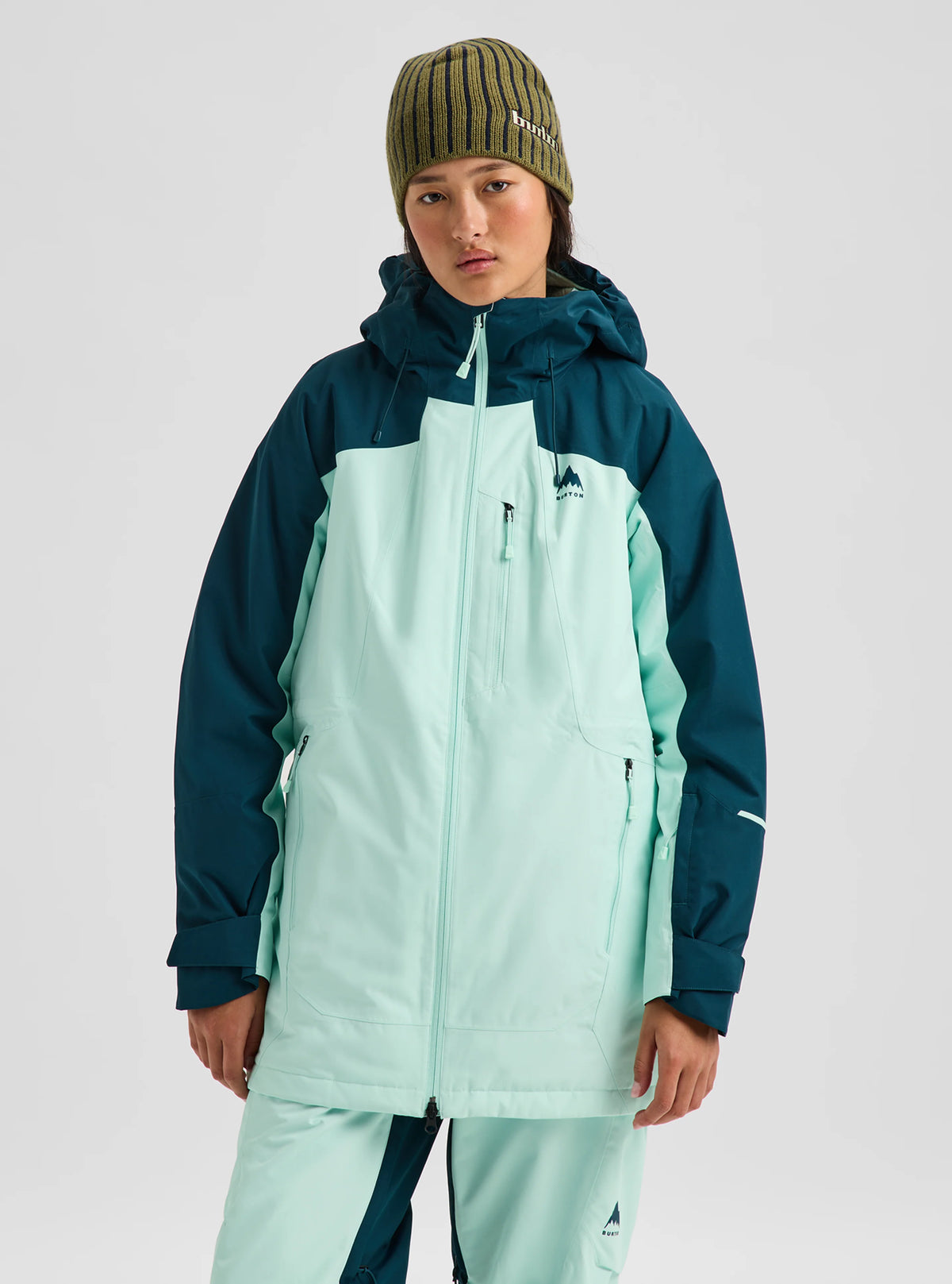 A model wearing a color-blocked snowboard jacket in mint green and dark teal, paired with matching pants, a striped beanie, and a face mask, against a light gray studio background.