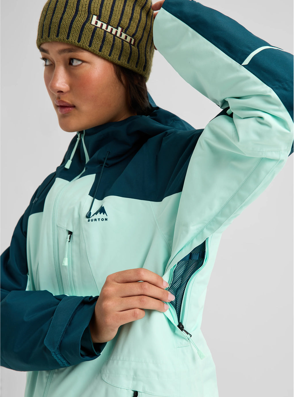 A model wearing a Burton ski jacket in mint green and dark teal, paired with a matching olive green striped beanie, against a light gray studio background.