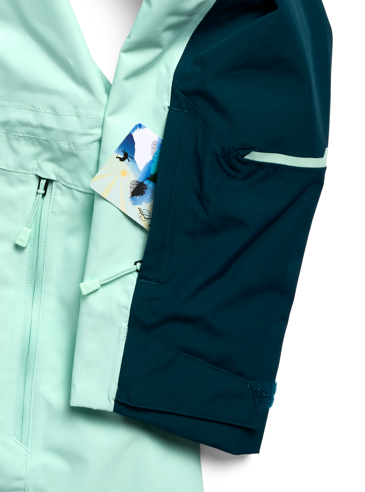 Close-up of a technical jacket with a split color design: mint green on the front and dark teal on the sleeve, featuring a zippered pocket and a small colorful card tucked into the pocket.