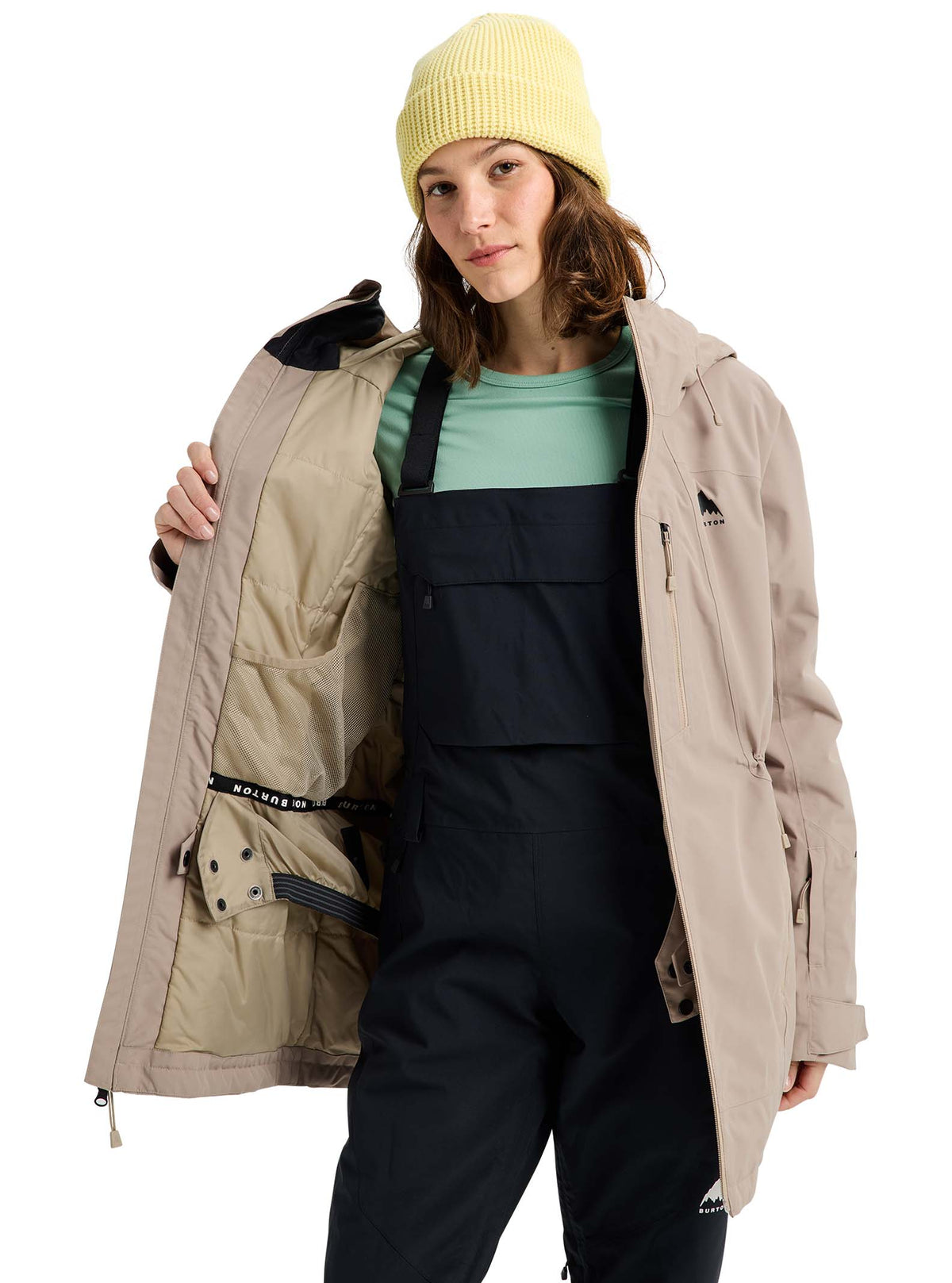 Person wearing a beige jacket and black bib overalls on a white background