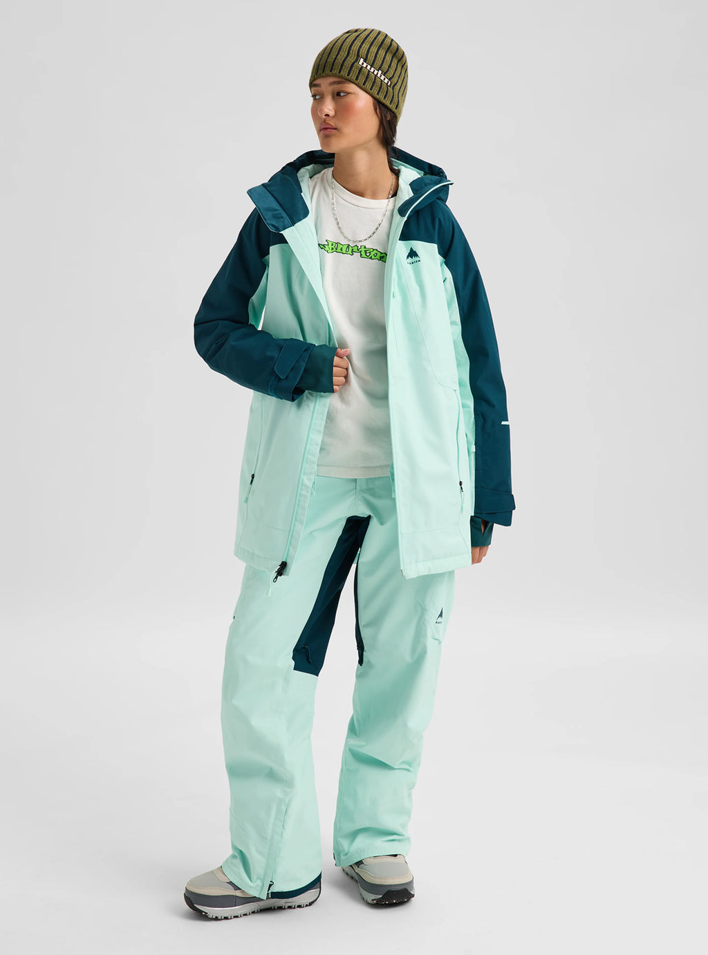 A model wearing a light mint green and dark teal snowboard jacket and matching pants, styled with a white graphic t-shirt, gray sneakers, and a green striped beanie against a light gray studio background.