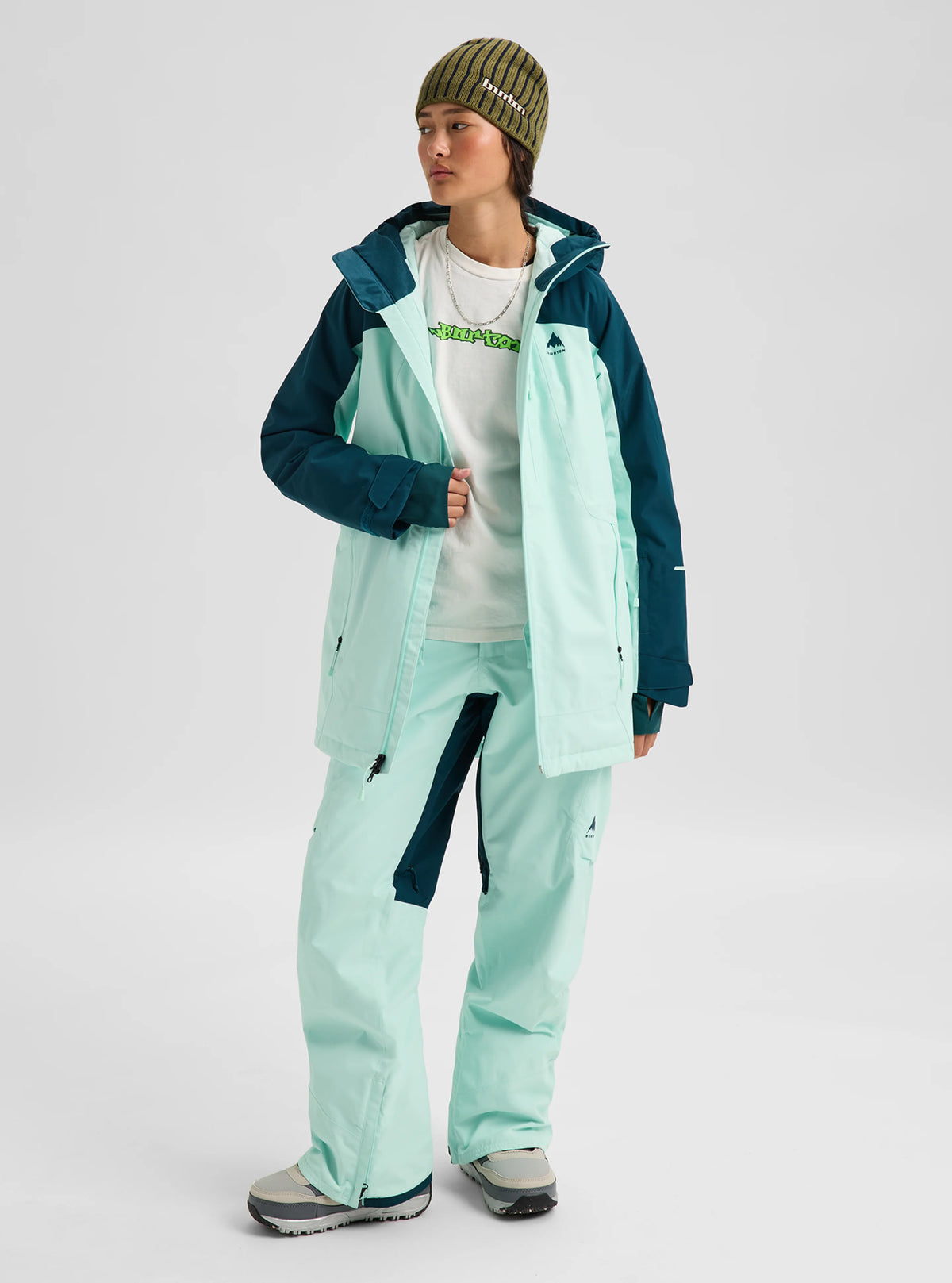 A model wearing a light mint green and dark teal snowboard jacket and matching pants, styled with a white graphic t-shirt, gray sneakers, and a green striped beanie against a light gray studio background.