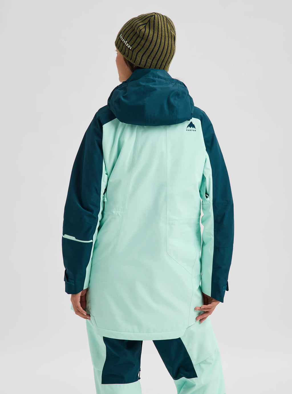 A person shown from the back wearing a mint green and dark teal snowboard jacket and matching snowboard pants, with a striped beanie, against a light gray studio background.