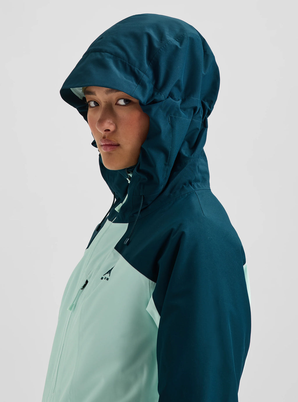 A model wearing a hooded technical jacket in dark teal and mint green, shown in a side profile against a light gray studio background.