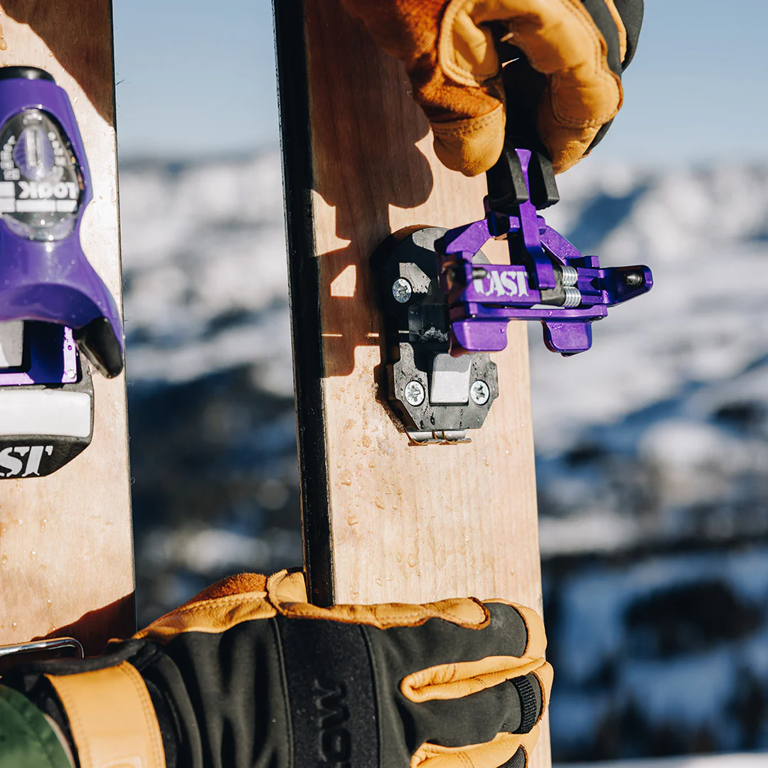 Close-up of a ski with purple bindings held by gloved hands against a blurred snowy background.