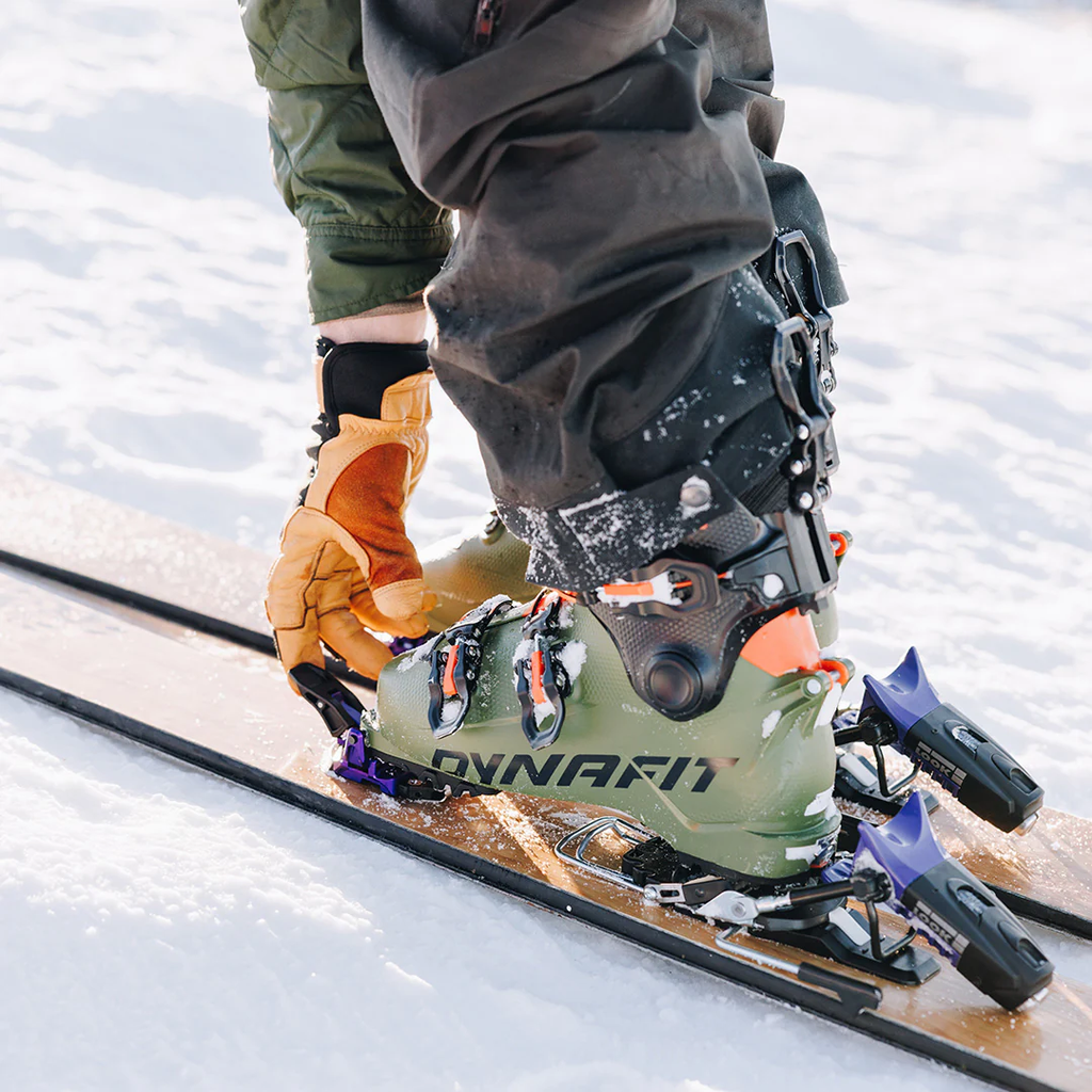 Close-up of a person's feet in green Dynafit ski boots attached to wooden skis on snow.