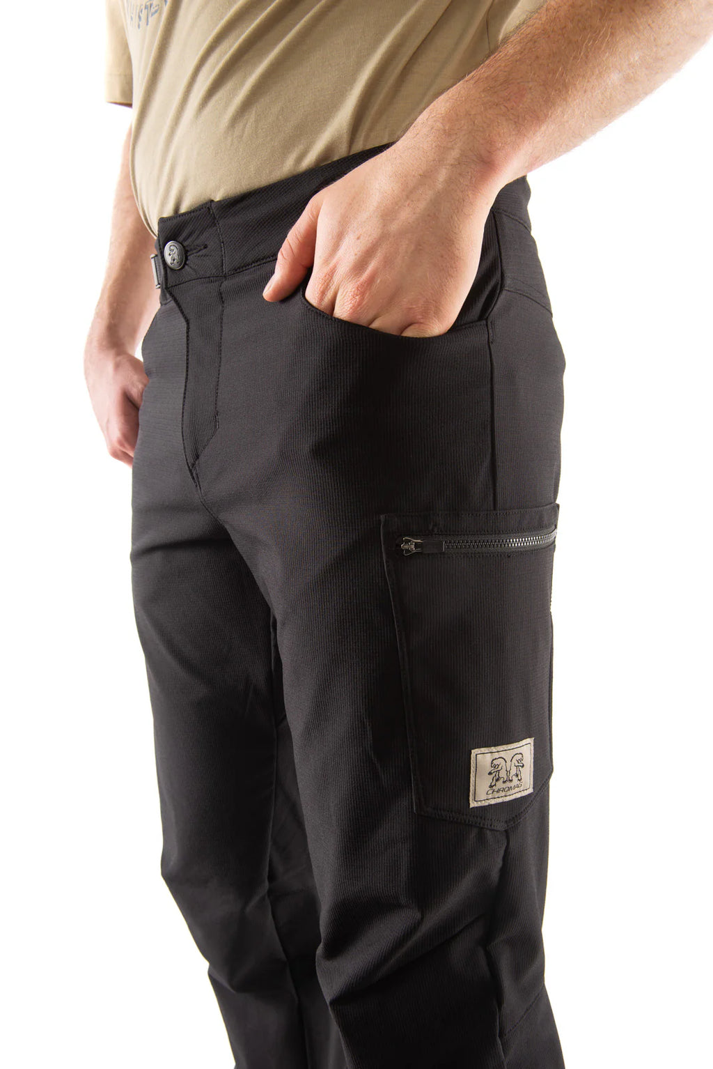 Close-up of a person wearing black cargo pants with a zippered side pocket and a small brand patch, paired with a tan shirt against a white background.