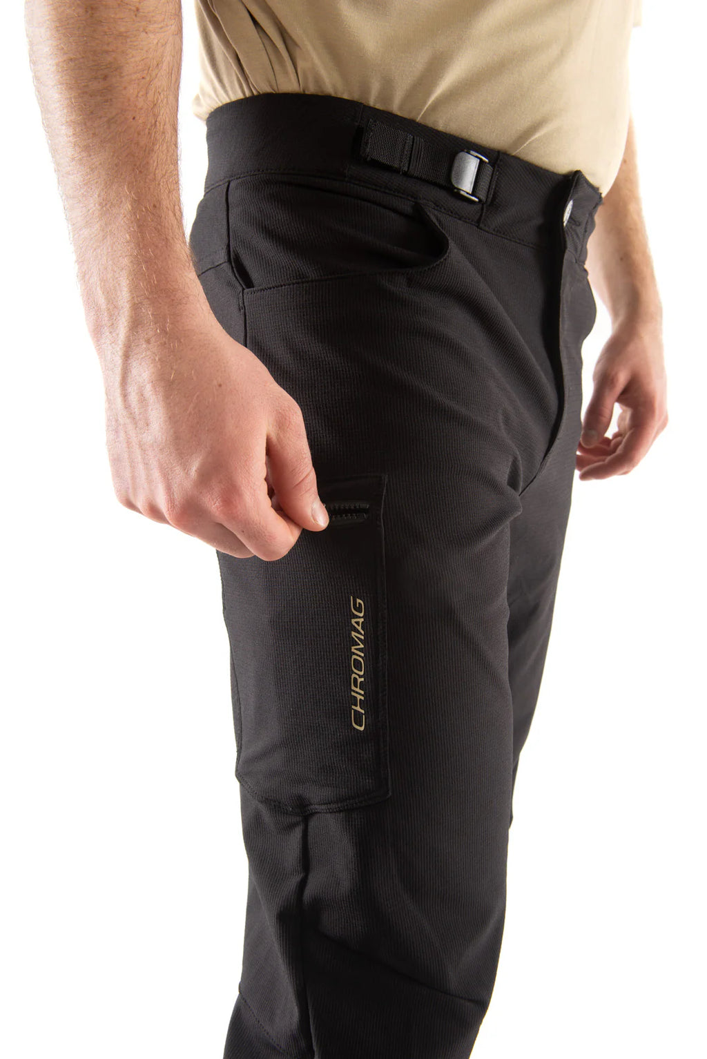 Close-up of a person wearing black technical pants with a vertical CHROMAGE logo on the side pocket, paired with a beige shirt, against a white background.