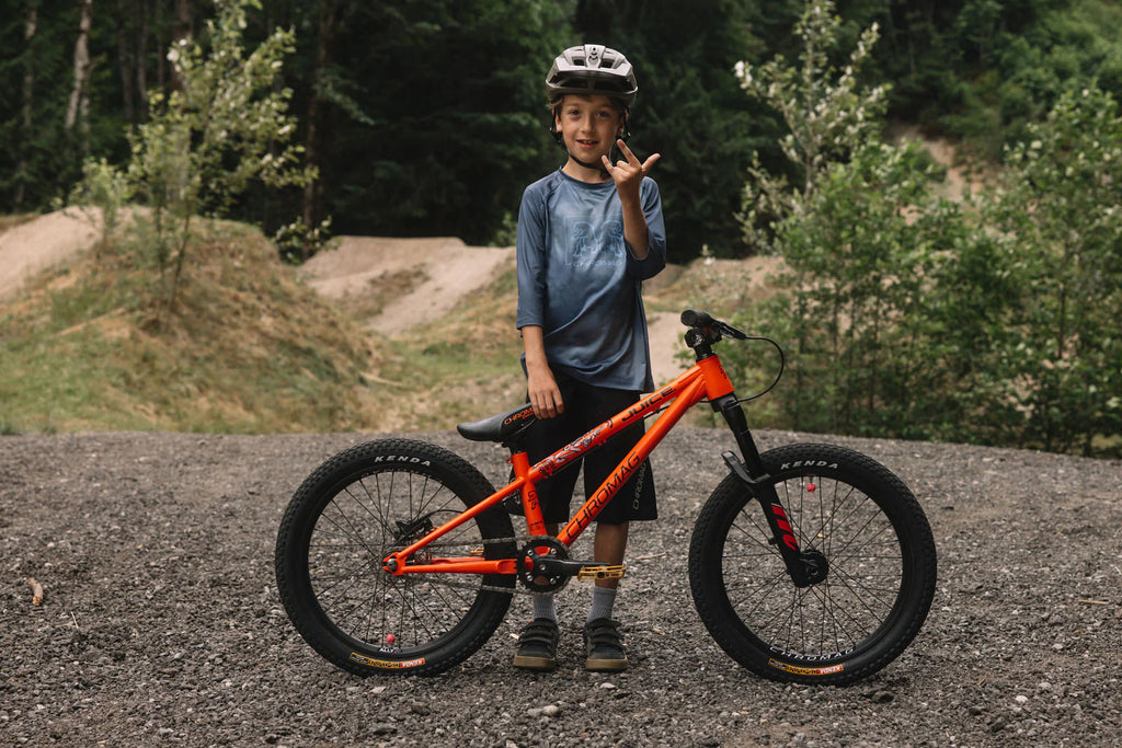 A child wearing a helmet and blue shirt stands next to an orange full-suspension mountain bike on a gravel path with a forest and dirt trail in the background.