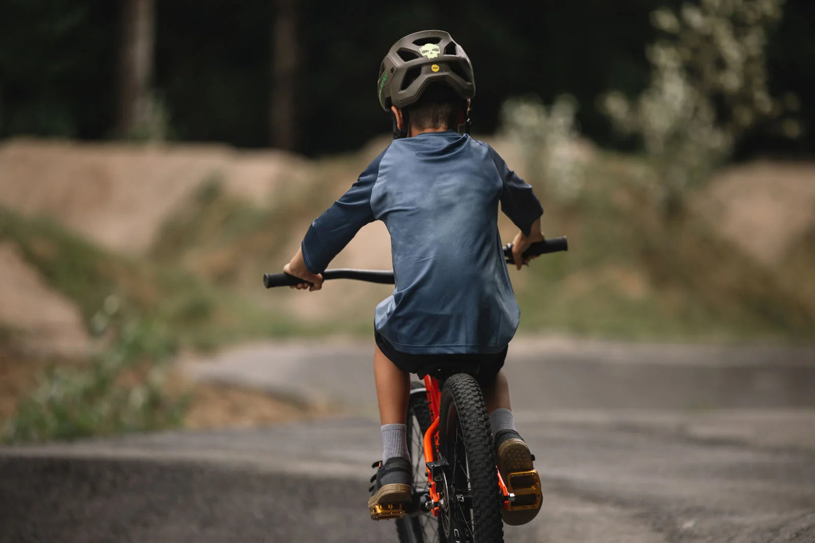 A child riding a bicycle on a paved path outdoors, viewed from behind. The rider wears a dark helmet, a blue long-sleeve shirt, dark shorts, and cycling shoes, with the bicycle visible in the foreground.