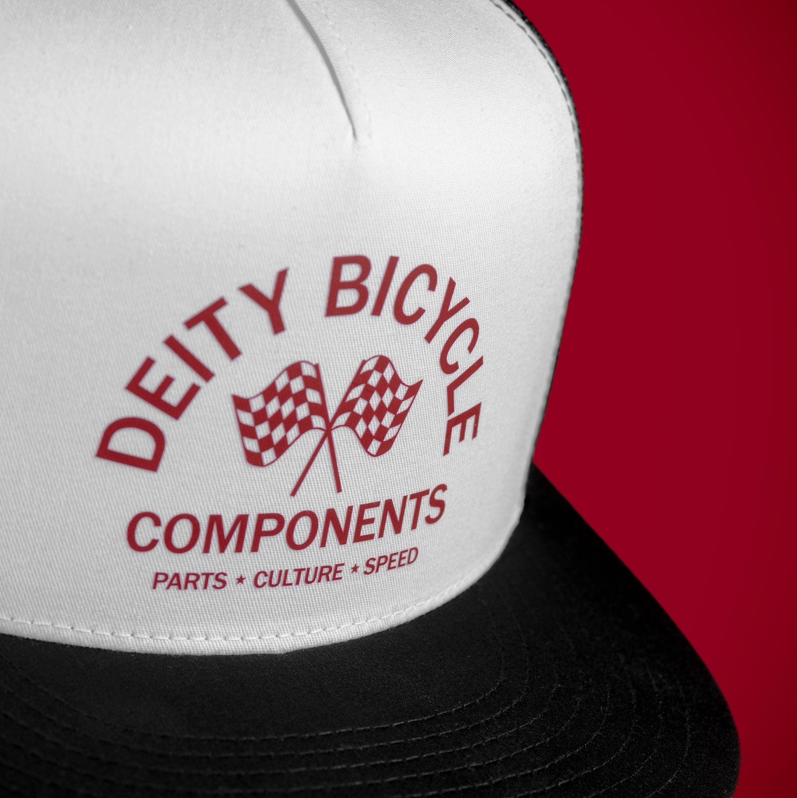 A close-up of a white and black trucker hat with red printed text reading 'DEITY BICYCLE COMPONENTS' and a checkered flag graphic, set against a solid red background.