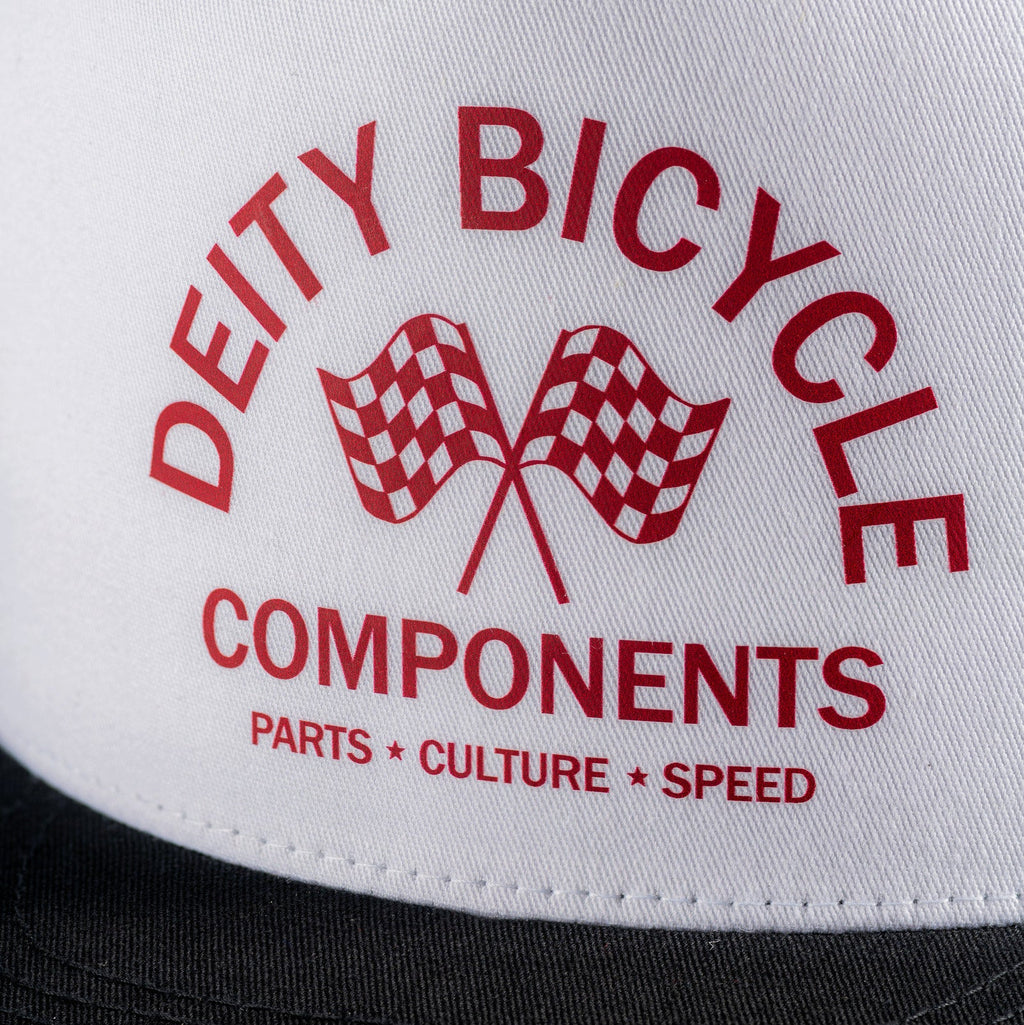 Close-up of a white trucker cap with red printed text reading 'DEITY BICYCLE COMPONENTS' and 'PARTS * CULTURE * SPEED,' plus two checkered flags, with a black brim visible at the bottom.