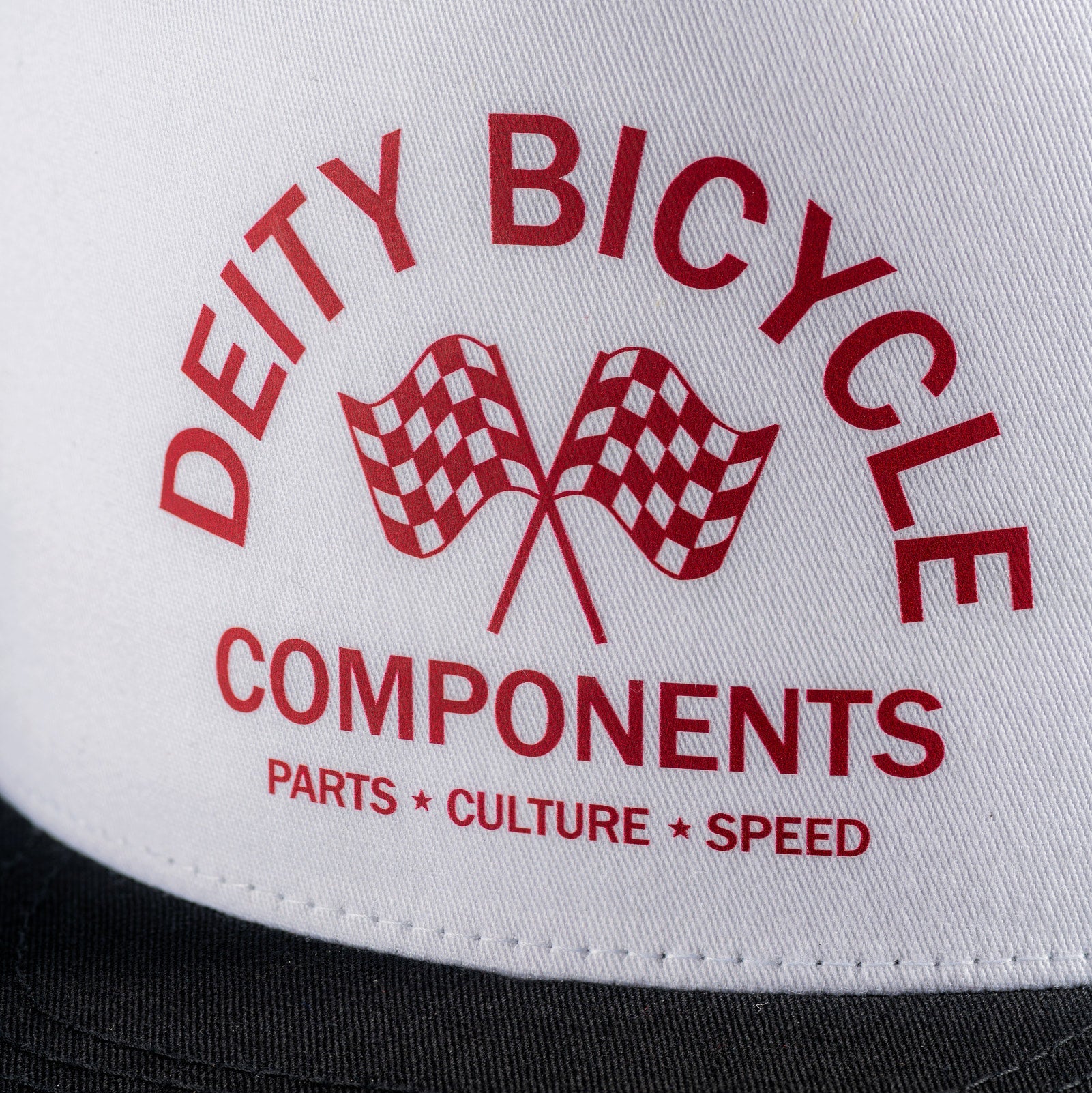 Close-up of a white trucker cap with red printed text reading 'DEITY BICYCLE COMPONENTS' and 'PARTS * CULTURE * SPEED,' plus two checkered flags, with a black brim visible at the bottom.