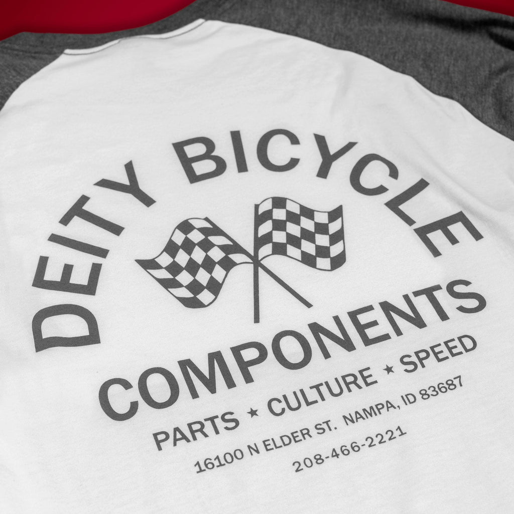 A close-up of a raglan baseball t-shirt showing the back graphic. The shirt has a white body and dark gray sleeves, featuring a black graphic print that reads 'DEITY BICYCLE COMPONENTS' with two checkered flags and contact details.