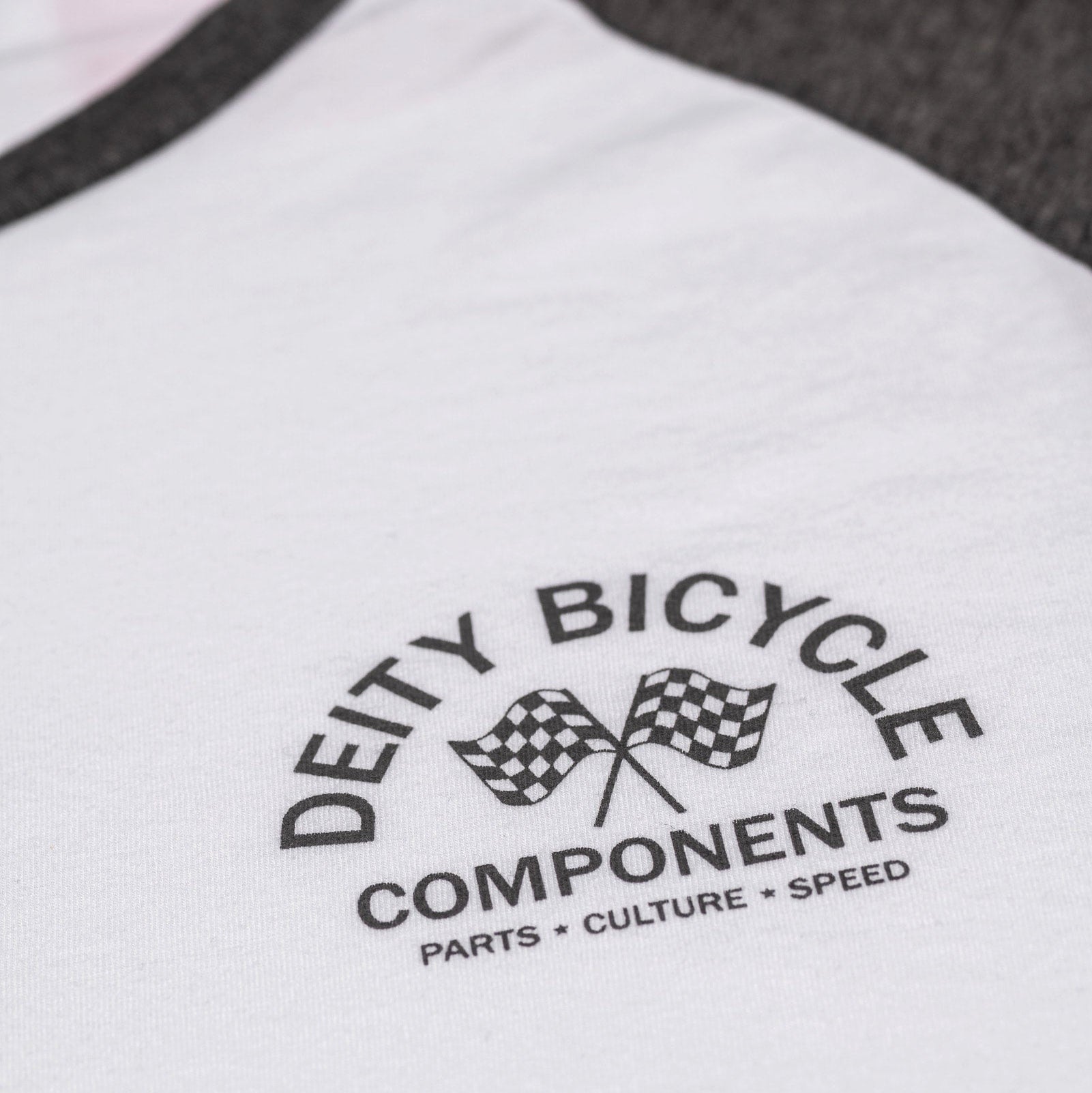 Close-up of a raglan baseball-style shirt with a white body, dark gray sleeves and collar trim, and a black printed chest logo reading 'DEITY BICYCLE COMPONENTS' with checkered flags and smaller text 'PARTS • CULTURE • SPEED'.