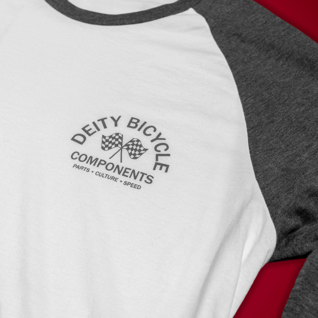 A close-up of a raglan baseball t-shirt with a white body and dark gray sleeves, featuring a black 'Deity Bicycle Components' logo printed on the chest, laid flat on a red background.