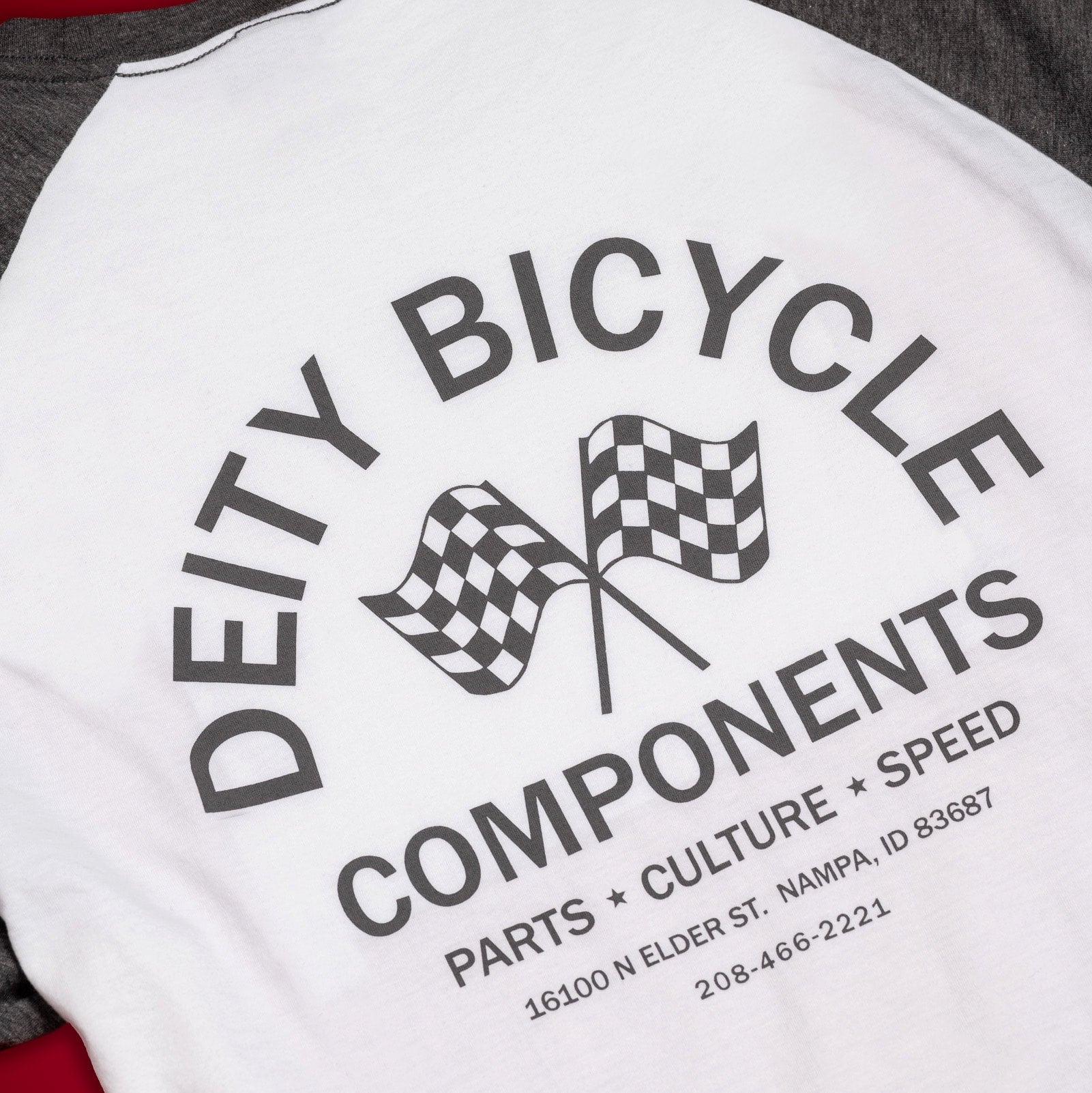 A close-up of a raglan-style baseball t-shirt showing the back graphic. The shirt has a white body and dark gray sleeves, featuring a black graphic print that reads 'DEITY BICYCLE COMPONENTS' with two checkered flags in the center, laid flat on a red background.
