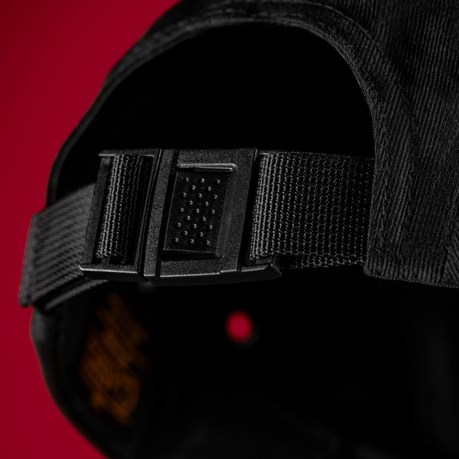 Close-up of a black baseball cap showing the adjustable rear strap and black plastic buckle, set against a solid red background.