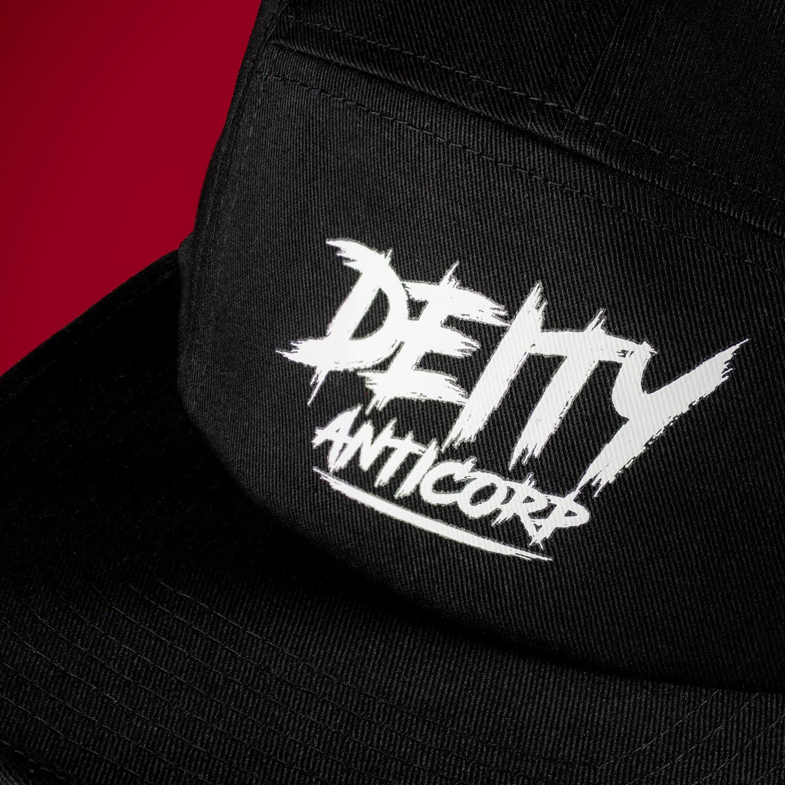 Close-up of a black snapback cap with a white embroidered 'DEITY ANTICORP' logo on the front panel, set against a solid red background.