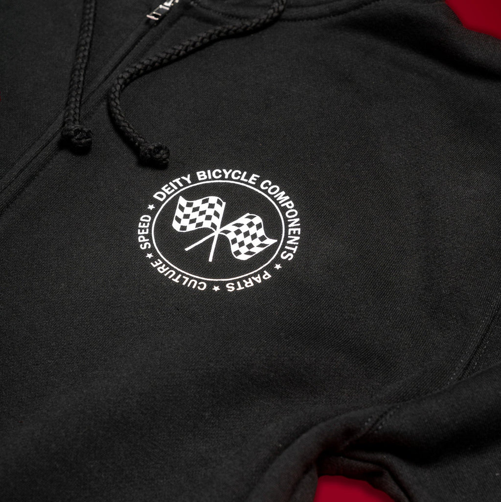 Close-up of a black hoodie with a white circular chest graphic reading 'Deity Bicycle Components' around two checkered flags, shown against a dark background with a red area visible at the lower right.