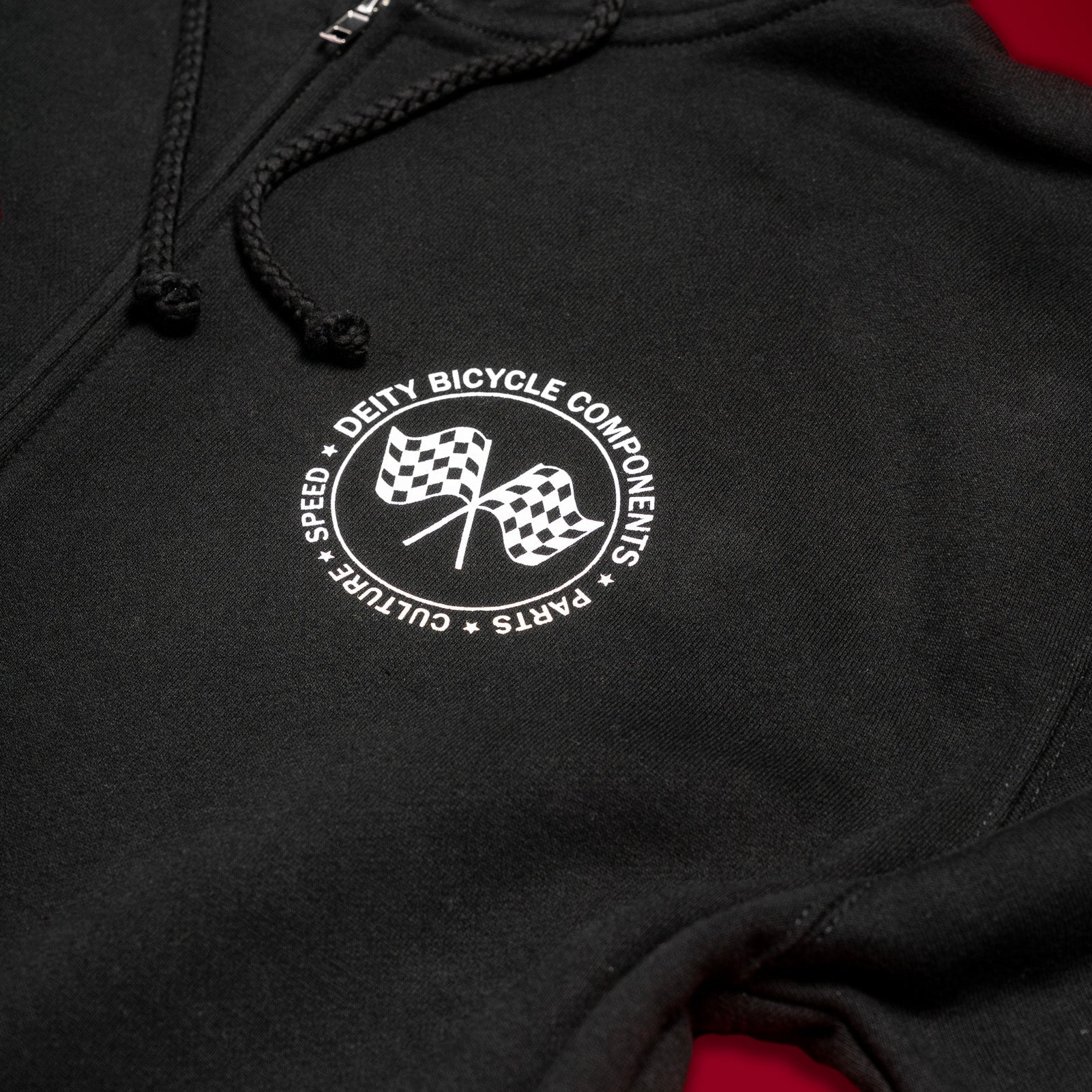 Close-up of a black hoodie with a white circular chest graphic reading 'Deity Bicycle Components' around two checkered flags, shown against a dark background with a red area visible at the lower right.