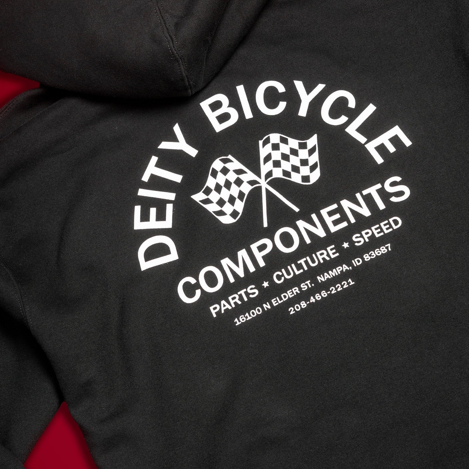 Close-up of a black hoodie showing the back graphic with white text reading 'DEITY BICYCLE COMPONENTS' and two checkered flags, with red fabric visible along the sides.