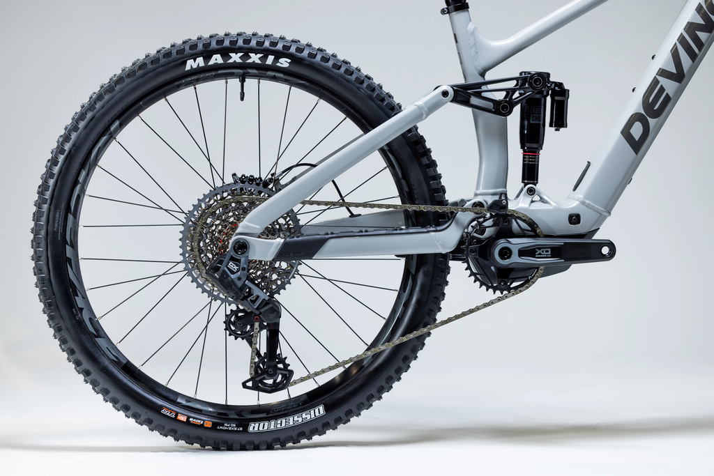Close-up of a mountain bike with a focus on the wheel and chain, featuring brand names Maxxis and Devinci.