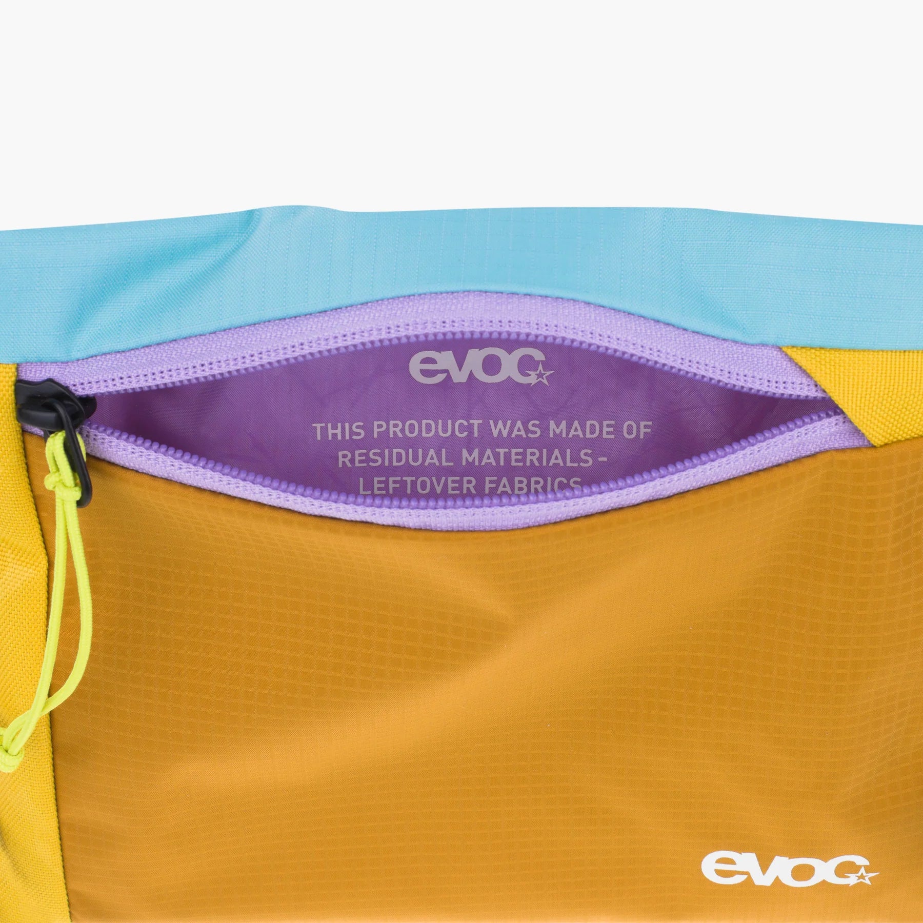 Close-up of a colorful zippered pouch showing a purple interior label that reads 'evoc' and 'THIS PRODUCT WAS MADE OF RESIDUAL MATERIALS - LEFTOVER FABRICS'. The pouch has a yellow-orange body, a light blue top section, and a yellow side panel, with a neon yellow zipper pull.