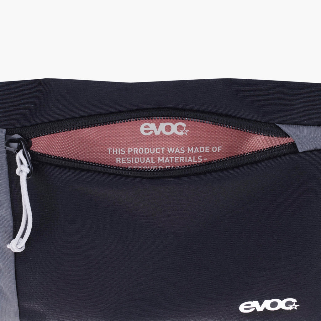 Close-up of a black EVOC bag showing a zipper opening with a dusty rose interior label that reads 'THIS PRODUCT WAS MADE OF RESIDUAL MATERIALS' and an EVOC logo, with a small white logo near the bottom right.