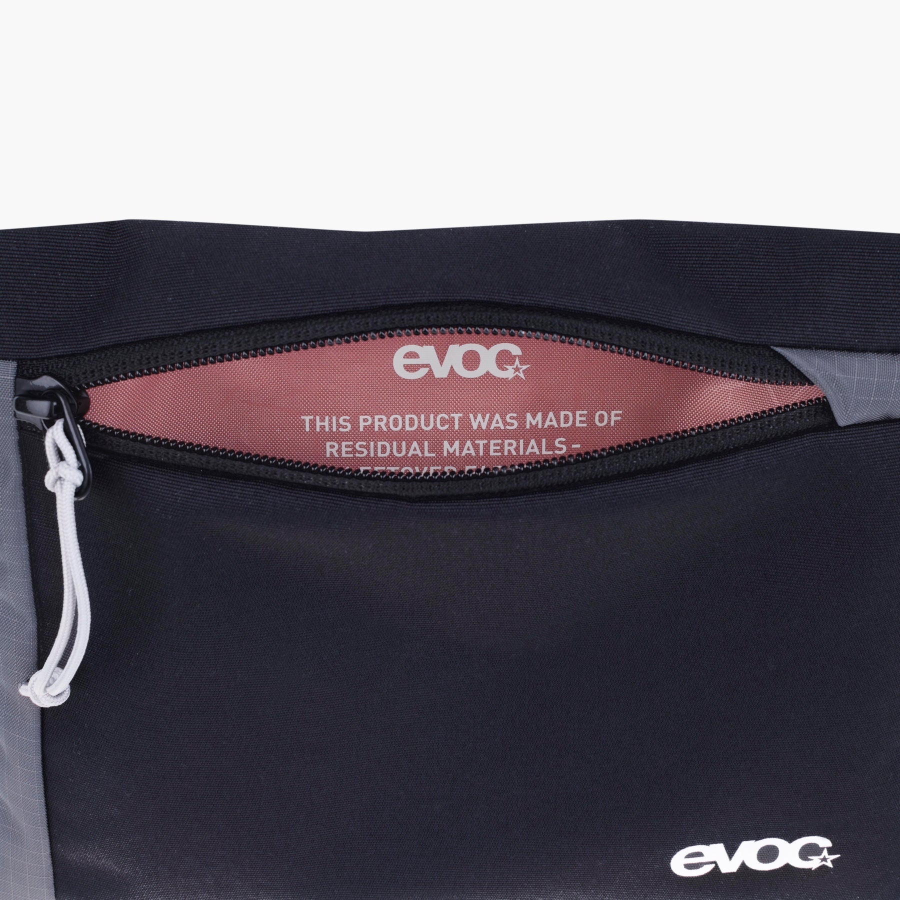 Close-up of a black EVOC bag showing a zipper opening with a dusty rose interior label that reads 'THIS PRODUCT WAS MADE OF RESIDUAL MATERIALS' and an EVOC logo, with a small white logo near the bottom right.