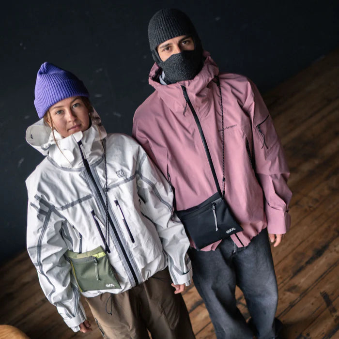 Two models wearing outdoor jackets and crossbody bags from the brand O'Neill, standing on a wooden floor against a dark background. One model wears a light gray jacket with a purple beanie and an olive green bag, while the other wears a dusty pink jacket with a black beanie, black face covering, and a black bag.