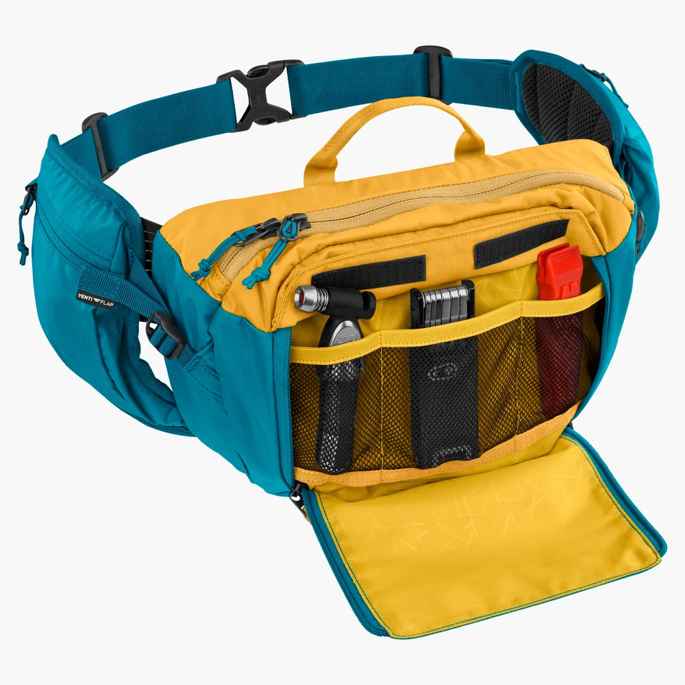 A teal and yellow waist bag shown open against a light gray background, with multiple zip compartments, mesh pockets, and small accessories stored inside.