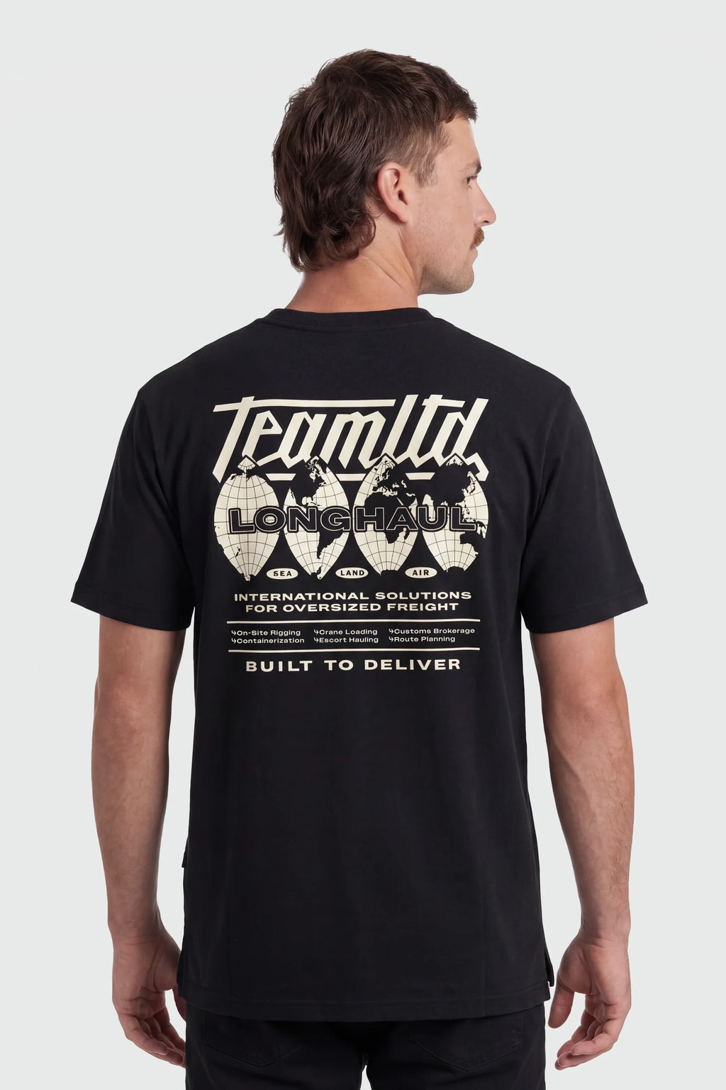 A man wearing a black short-sleeve t-shirt, viewed from the back. The shirt features a large white graphic print on the back with the text 'Team Ltd', 'LONG Haul', and 'Built to Deliver' along with globe illustrations.