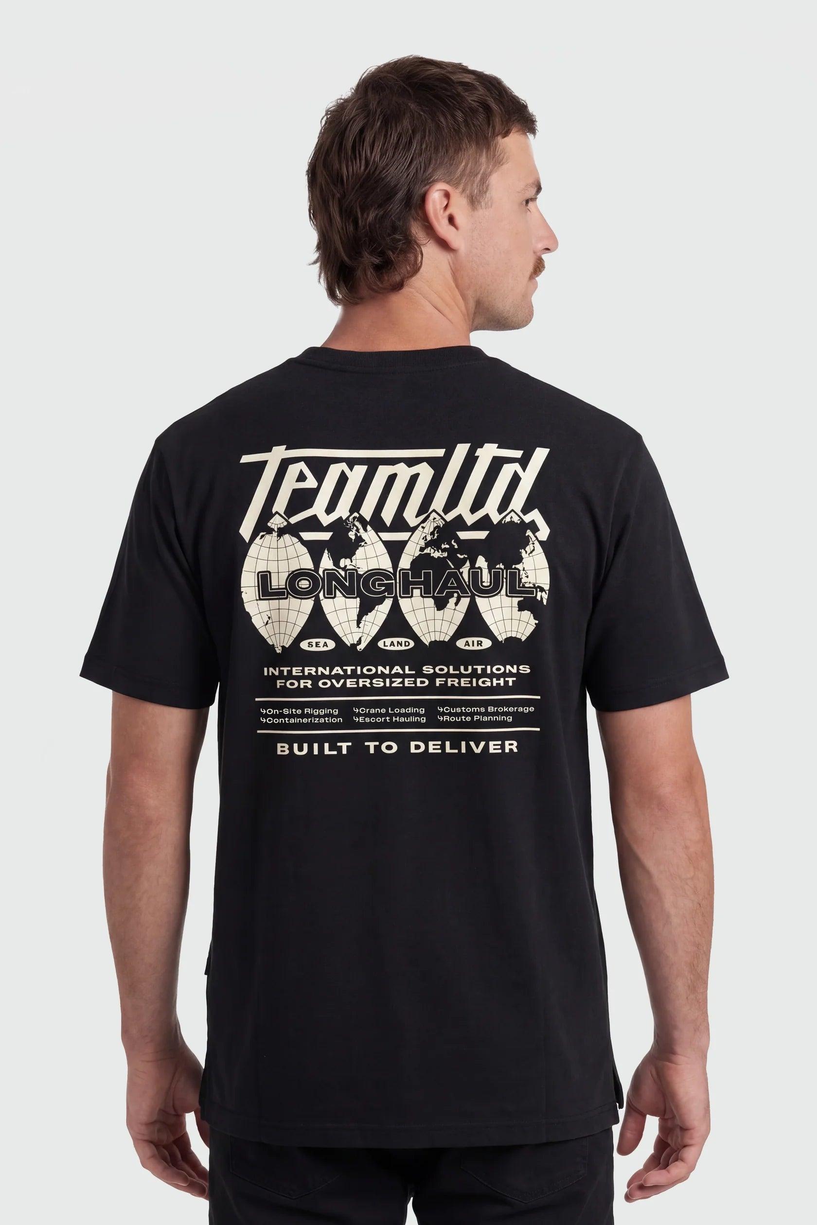 A man wearing a black short-sleeve t-shirt, viewed from the back. The shirt features a large white graphic print on the back with the text 'Team Ltd', 'LONG Haul', and 'Built to Deliver' along with globe illustrations.