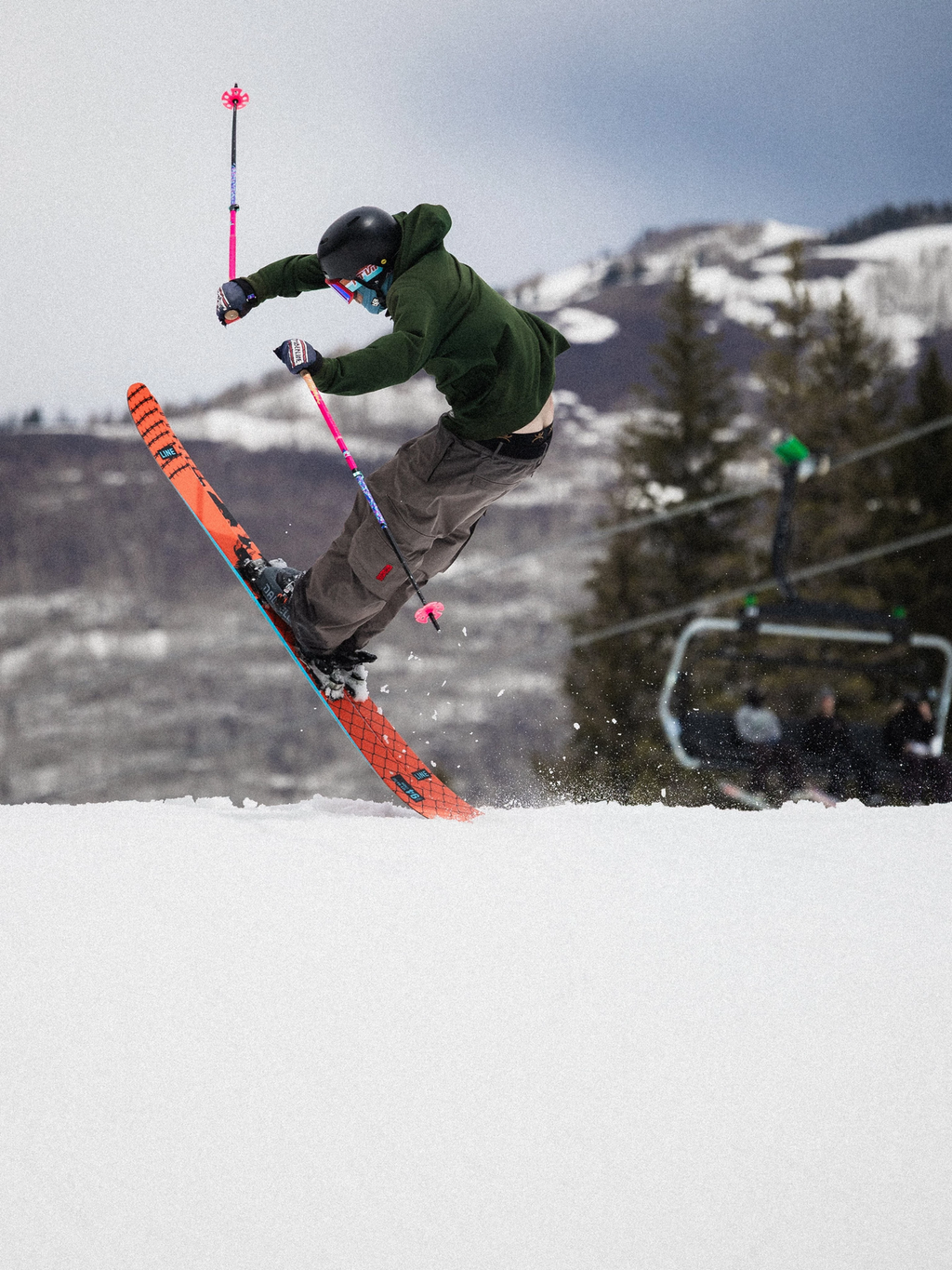 Skier performing a trick in the air with a ski lift and snowy mountains in the background