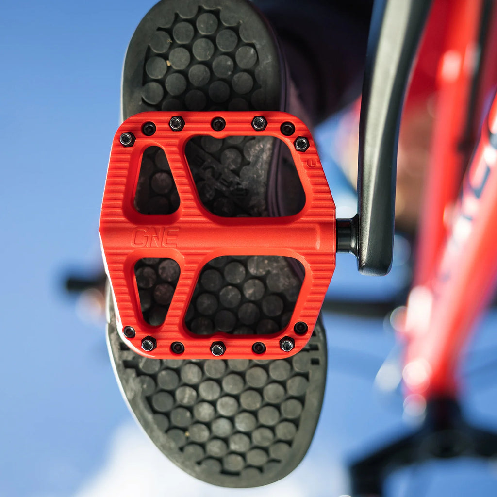 A close-up shot of a bright red bicycle pedal with black cleats, shown from a low angle against a blue sky and blurred red bicycle frame.