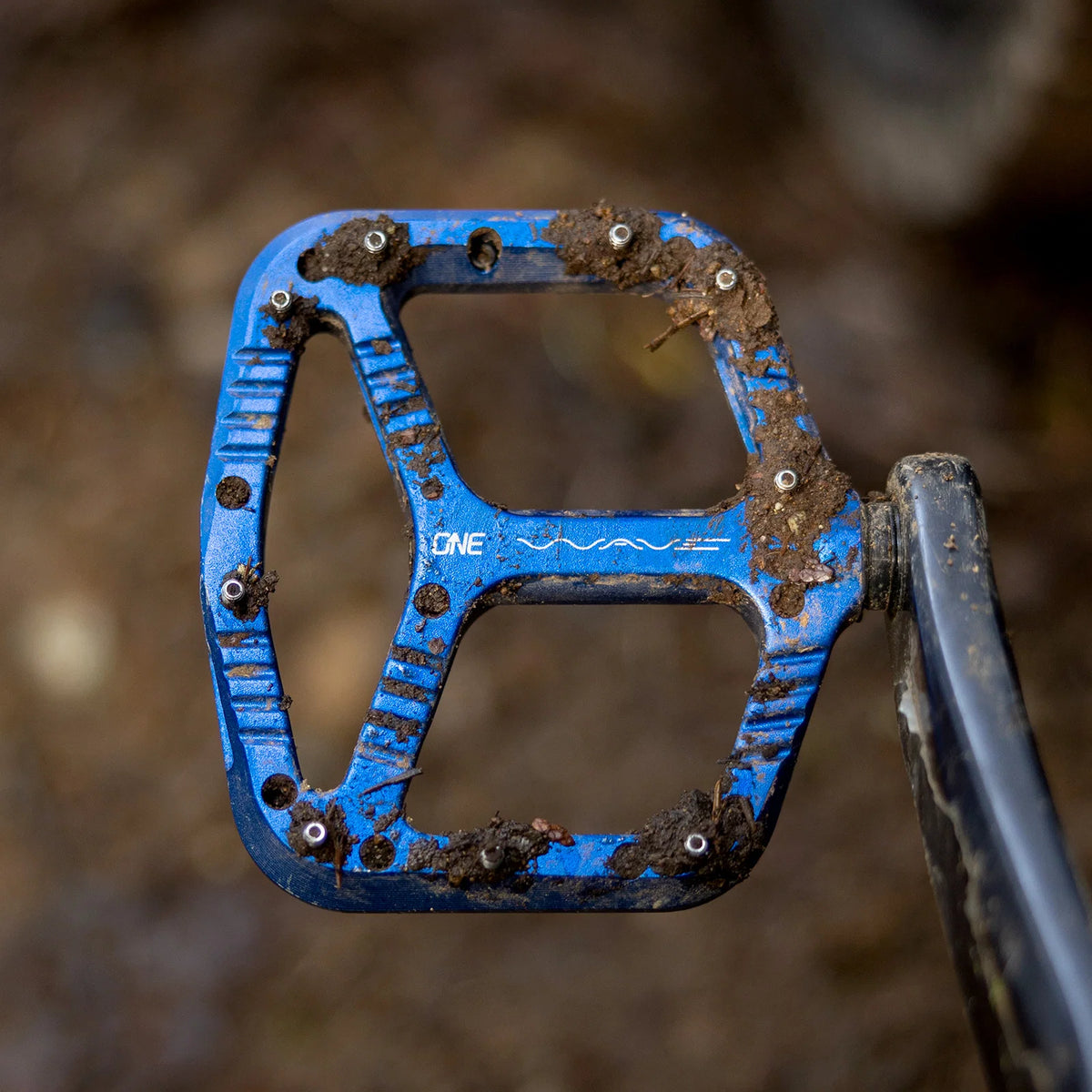 A close-up of a blue mountain bike pedal covered in dirt and mud, with the brand name 'ONE' visible on the platform.