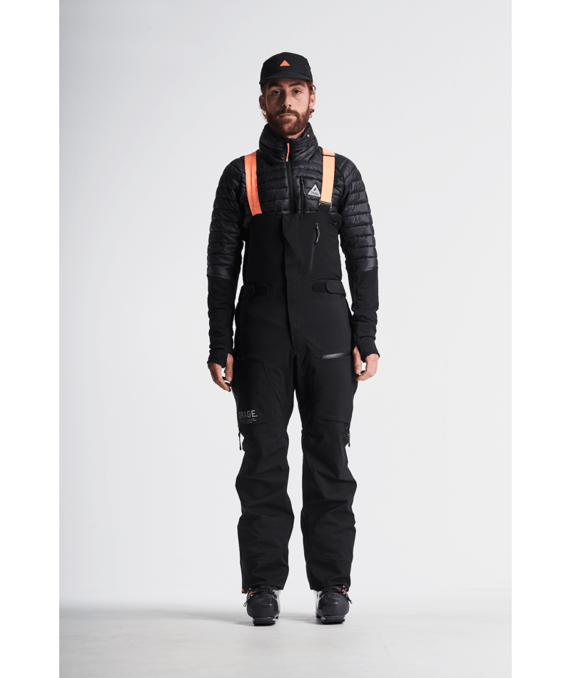 Person wearing a black snowsuit with orange suspenders on a white background