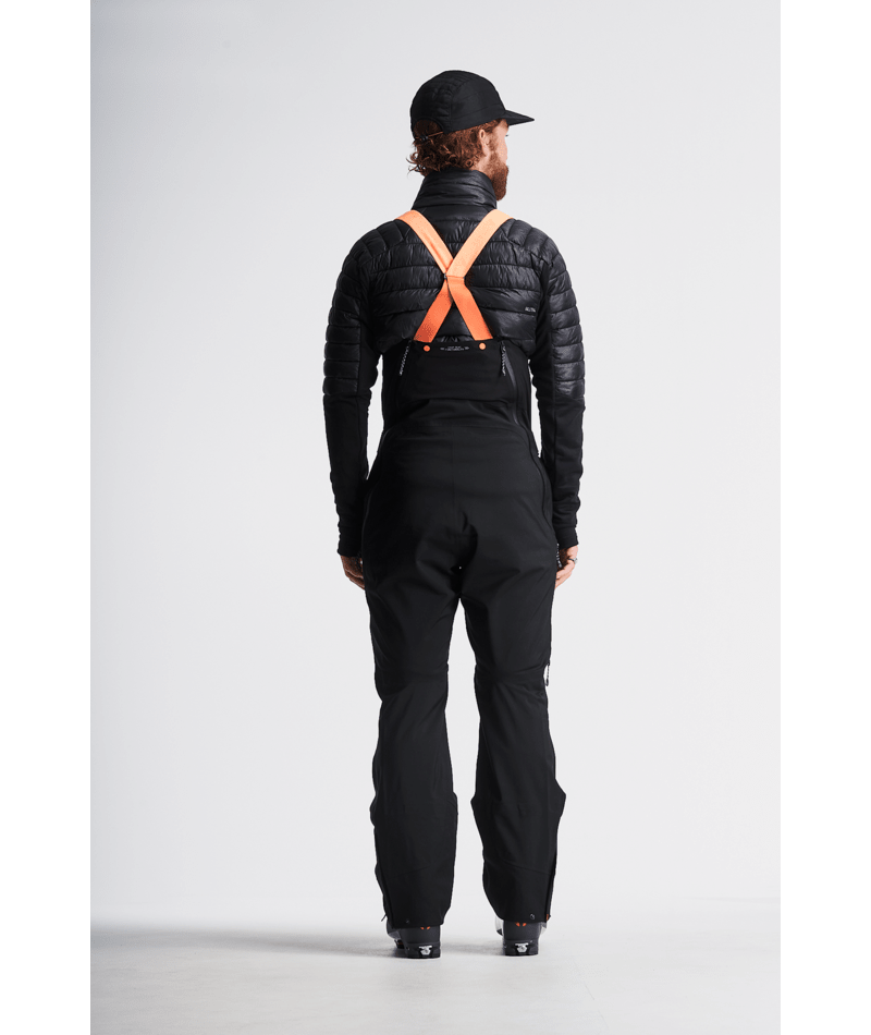 Person wearing a black puffer jacket and black bib overalls with orange suspenders on a light gray background
